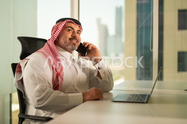 إدارة شؤون الأعمال وتنظيمها، استخدام جهاز تقني حديث ومتطور، مهنة ووظيفة ادارية مكتبية، رجل عربي خليجي سعودي يرتدي الثوب التقليدي والشماغ يقوم بالعمل على الحاسوب المحمول داخل المكتب، الشركات والمكاتب في المملكة العربية السعودية، اجراء مكالمة هاتفية