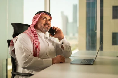 إدارة شؤون الأعمال وتنظيمها، استخدام جهاز تقني حديث ومتطور، مهنة ووظيفة ادارية مكتبية، رجل عربي خليجي سعودي يرتدي الثوب التقليدي والشماغ يقوم بالعمل على الحاسوب المحمول داخل المكتب، الشركات والمكاتب في المملكة العربية السعودية، اجراء مكالمة هاتفية