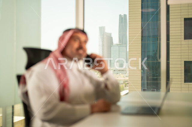 إدارة شؤون الأعمال وتنظيمها، استخدام جهاز تقني حديث ومتطور، مهنة ووظيفة ادارية مكتبية، رجل عربي خليجي سعودي يرتدي الثوب التقليدي والشماغ يقوم بالعمل على الحاسوب المحمول داخل المكتب، الشركات والمكاتب في المملكة العربية السعودية، اجراء مكالمة هاتفية