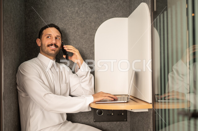 اجراء مكالمة عبر الهاتف المحمول, إدارة شؤون الأعمال وتنظيمها، استخدام جهاز تقني حديث ومتطور، مهنة ووظيفة ادارية مكتبية، رجل عربي خليجي سعودي يرتدي الثوب التقليدي يقوم بالعمل على الحاسوب المحمول داخل المكتب، الشركات والمكاتب في المملكة العربية السعودية