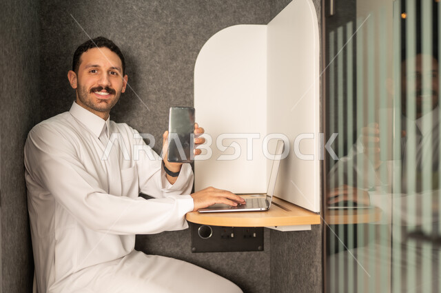 عرض شاشة هاتف محمول سوداء فارغة, إدارة شؤون الأعمال وتنظيمها، استخدام جهاز تقني حديث ومتطور، مهنة ووظيفة ادارية مكتبية، رجل عربي خليجي سعودي يرتدي الثوب التقليدي يقوم بالعمل على الحاسوب المحمول داخل المكتب، الشركات والمكاتب في المملكة العربية السعودية