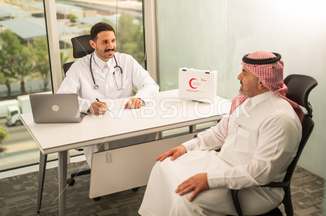  تسجيل حالات المرضى على جهاز الحاسوب, طبيب عربي خليجي سعودي يرتدي المعطف الأبيض يقوم بشرح الحالة المرضية لمريض يرتدي الشماغ والثوب التقليدي, الحوار مع المرضى لكشف الحالة، تقديم الاستشارات في المراكز الطبيه, متابعة السجل الطبي للمراجعين, العمل في القطاعات الصحية