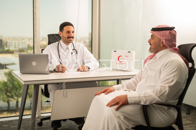  تسجيل حالات المرضى على جهاز الحاسوب, طبيب عربي خليجي سعودي يرتدي المعطف الأبيض يقوم بشرح الحالة المرضية لمريض يرتدي الشماغ والثوب التقليدي, الحوار مع المرضى لكشف الحالة، تقديم الاستشارات في المراكز الطبيه, متابعة السجل الطبي للمراجعين, العمل في القطاعات الصحية
