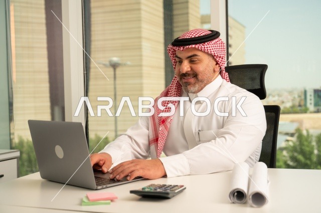 إدارة شؤون الأعمال وتنظيمها، استخدام جهاز تقني حديث ومتطور، مهنة ووظيفة ادارية مكتبية، موظف عربي خليجي سعودي يرتدي الثوب والشماغ التقليدي يقوم بالعمل على الحاسوب المحمول داخل المكتب، الشركات والمكاتب في المملكة العربية السعودية