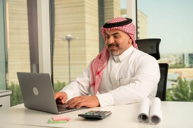 إدارة شؤون الأعمال وتنظيمها، استخدام جهاز تقني حديث ومتطور، مهنة ووظيفة ادارية مكتبية، موظف عربي خليجي سعودي يرتدي الثوب والشماغ التقليدي يقوم بالعمل على الحاسوب المحمول داخل المكتب، الشركات والمكاتب في المملكة العربية السعودية