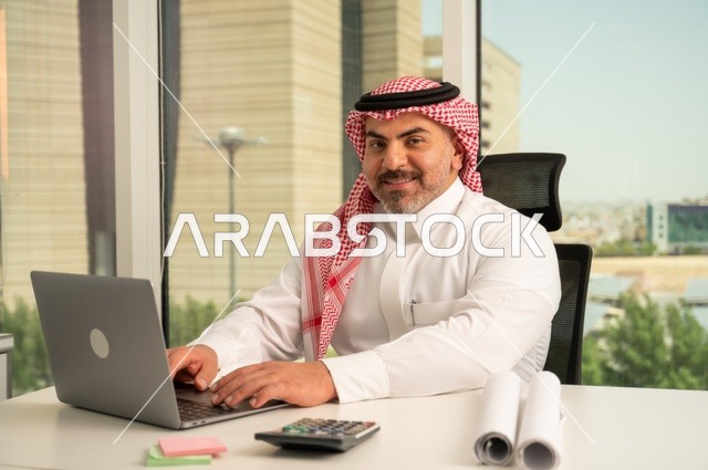Looking at the camera with expressions of joy, an administrative office profession and job, managing and organizing business affairs, using a modern and advanced technical device, an Arab Gulf Saudi employee wearing traditional thobe and ghutrah working on a laptop inside the office, companies and offices in the Kingdom of Saudi Arabia.