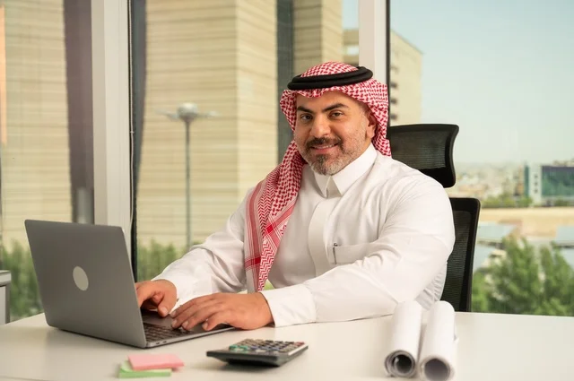 النظر إلى الكاميرا بتعابير السرور، مهنة ووظيفة ادارية مكتبية، إدارة شؤون الأعمال وتنظيمها، استخدام جهاز تقني حديث ومتطور، موظف عربي خليجي سعودي يرتدي الثوب والشماغ التقليدي يقوم بالعمل على الحاسوب المحمول داخل المكتب، الشركات والمكاتب في المملكة العربية السعودية