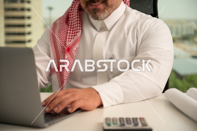 A profession and administrative office job, managing and organizing business affairs, using modern and advanced technology, a close-up shot of a Saudi Arabian Gulf Arab employee wearing traditional thobe and ghutrah working on a laptop inside the office, companies and offices in the Kingdom of Saudi Arabia.