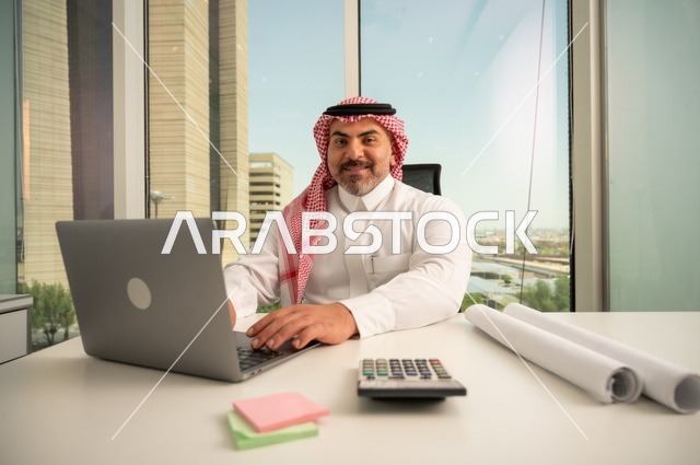 Looking at the camera with expressions of joy, an administrative office profession and job, managing and organizing business affairs, using a modern and advanced technical device, an Arab Gulf Saudi employee wearing traditional thobe and ghutrah working on a laptop inside the office, companies and offices in the Kingdom of Saudi Arabia.