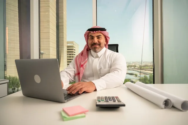 النظر إلى الكاميرا بتعابير السرور، مهنة ووظيفة ادارية مكتبية، إدارة شؤون الأعمال وتنظيمها، استخدام جهاز تقني حديث ومتطور، موظف عربي خليجي سعودي يرتدي الثوب والشماغ التقليدي يقوم بالعمل على الحاسوب المحمول داخل المكتب، الشركات والمكاتب في المملكة العربية السعودية