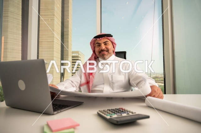 Working on a laptop inside the office, looking at the camera with expressions of joy, an administrative office profession and job, managing business affairs through a modern and advanced technical device, an Arab Gulf Saudi employee wearing traditional thobe and shemagh reading and reviewing plans, companies and offices in Saudi Arabia.