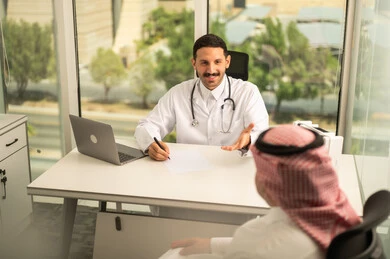 Recording patient cases on a computer, a Saudi Arabian Gulf doctor wearing a white coat explaining the medical condition of a patient wearing a traditional keffiyeh and thobe, dialogue with patients to determine the condition, providing consultations in medical centers, following up on the medical records of patients, working in the health sector