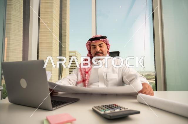 إيماءات الاندماج والتركيز، العمل على الحاسوب المحمول داخل المكتب، مهنة ووظيفة ادارية مكتبية، إدارة شؤون الأعمال عبر جهاز تقني حديث ومتطور، موظف عربي خليجي سعودي يرتدي الثوب والشماغ التقليدي يقوم بقراءة وتدقيق المخططات، الشركات والمكاتب في السعودية