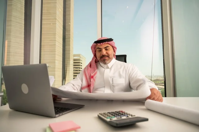 إيماءات الاندماج والتركيز، العمل على الحاسوب المحمول داخل المكتب، مهنة ووظيفة ادارية مكتبية، إدارة شؤون الأعمال عبر جهاز تقني حديث ومتطور، موظف عربي خليجي سعودي يرتدي الثوب والشماغ التقليدي يقوم بقراءة وتدقيق المخططات، الشركات والمكاتب في السعودية
