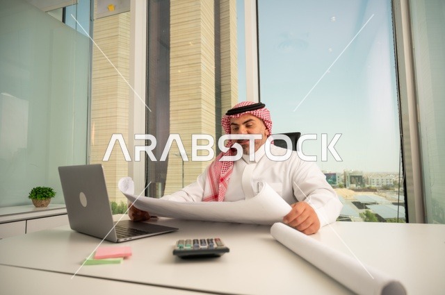 إيماءات الاندماج والتركيز، العمل على الحاسوب المحمول داخل المكتب، مهنة ووظيفة ادارية مكتبية، إدارة شؤون الأعمال عبر جهاز تقني حديث ومتطور، موظف عربي خليجي سعودي يرتدي الثوب والشماغ التقليدي يقوم بقراءة وتدقيق المخططات، الشركات والمكاتب في السعودية