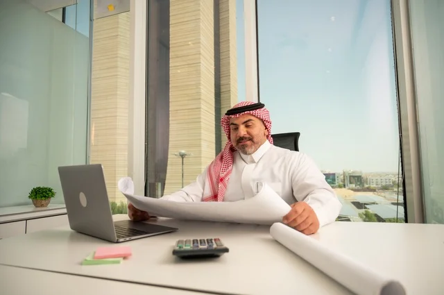 Gestures of integration and focus, working on a laptop inside the office, an administrative office profession and job, managing business affairs through a modern and advanced technical device, a Saudi Gulf Arab employee wearing the traditional thobe and ghutrah reading and reviewing plans, companies and offices in Saudi Arabia.