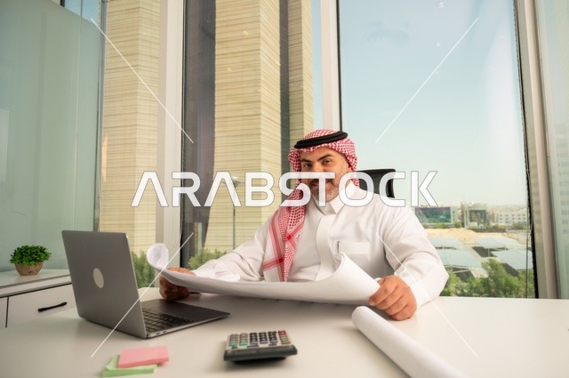 العمل على الحاسوب المحمول داخل المكتب، النظر إلى الكاميرا بتعابير السرور، مهنة ووظيفة ادارية مكتبية، إدارة شؤون الأعمال عبر جهاز تقني حديث ومتطور، موظف عربي خليجي سعودي يرتدي الثوب والشماغ التقليدي يقوم بقراءة وتدقيق المخططات، الشركات والمكاتب في السعودية