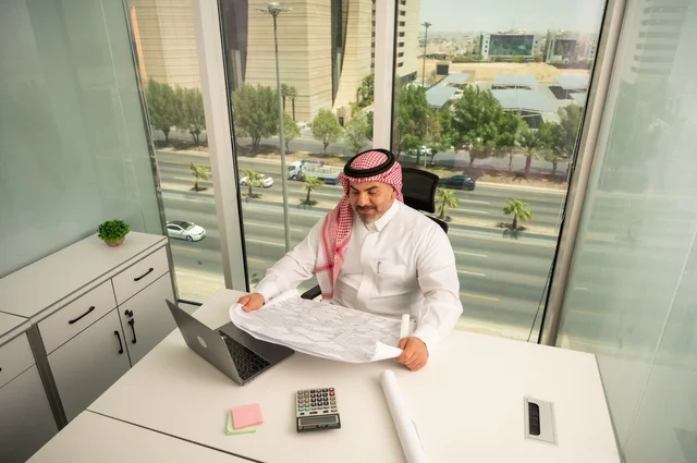 Gestures of integration and focus, working on a laptop inside the office, an administrative office profession and job, managing business affairs through a modern and advanced technical device, a Saudi Gulf Arab employee wearing the traditional thobe and ghutrah reading and reviewing plans, companies and offices in Saudi Arabia.