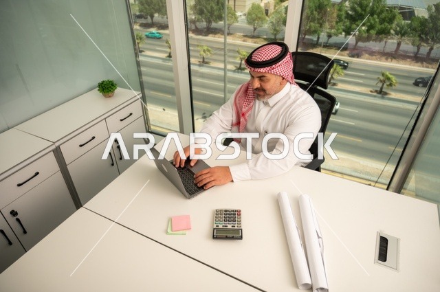 إدارة شؤون الأعمال وتنظيمها، استخدام جهاز تقني حديث ومتطور، مهنة ووظيفة ادارية مكتبية، موظف عربي خليجي سعودي يرتدي الثوب والشماغ التقليدي يقوم بالعمل على الحاسوب المحمول داخل المكتب، الشركات والمكاتب في المملكة العربية السعودية