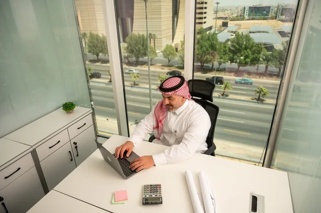 Management and organization of business affairs, using modern and advanced technology, an administrative office profession and job, an Arab Gulf Saudi employee wearing traditional thobe and ghutrah working on a laptop inside the office, companies and offices in the Kingdom of Saudi Arabia.