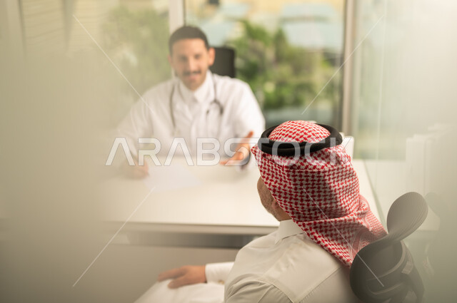  تسجيل حالات المرضى على جهاز الحاسوب, طبيب عربي خليجي سعودي يرتدي المعطف الأبيض يقوم بشرح الحالة المرضية لمريض يرتدي الشماغ والثوب التقليدي, الحوار مع المرضى لكشف الحالة، تقديم الاستشارات في المراكز الطبيه, متابعة السجل الطبي للمراجعين, العمل في القطاعات الصحية