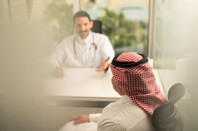Recording patient cases on a computer, a Saudi Arabian Gulf doctor wearing a white coat explaining the medical condition of a patient wearing a traditional keffiyeh and thobe, dialogue with patients to determine the condition, providing consultations in medical centers, following up on the medical records of patients, working in the health sector