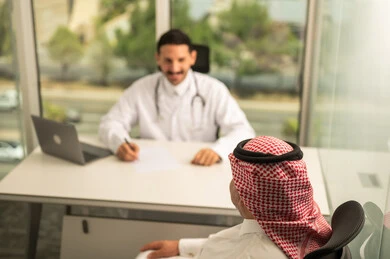 Recording patient cases on a computer, a Saudi Arabian Gulf doctor wearing a white coat explaining the medical condition of a patient wearing a traditional keffiyeh and thobe, dialogue with patients to determine the condition, providing consultations in medical centers, following up on the medical records of patients, working in the health sector