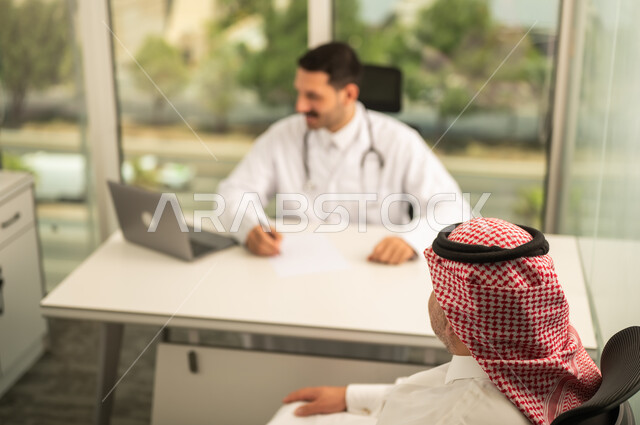  تسجيل حالات المرضى على جهاز الحاسوب, طبيب عربي خليجي سعودي يرتدي المعطف الأبيض يقوم بشرح الحالة المرضية لمريض يرتدي الشماغ والثوب التقليدي, الحوار مع المرضى لكشف الحالة، تقديم الاستشارات في المراكز الطبيه, متابعة السجل الطبي للمراجعين, العمل في القطاعات الصحية