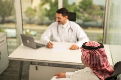 Recording patient cases on a computer, a Saudi Arabian Gulf doctor wearing a white coat explaining the medical condition of a patient wearing a traditional keffiyeh and thobe, dialogue with patients to determine the condition, providing consultations in medical centers, following up on the medical records of patients, working in the health sector