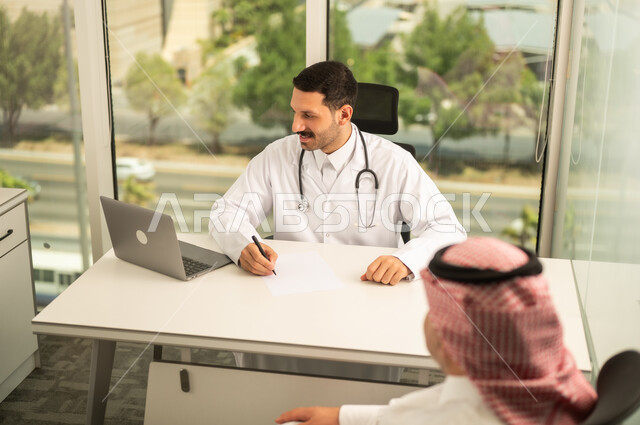  تسجيل حالات المرضى على جهاز الحاسوب, طبيب عربي خليجي سعودي يرتدي المعطف الأبيض يقوم بشرح الحالة المرضية لمريض يرتدي الشماغ والثوب التقليدي, الحوار مع المرضى لكشف الحالة، تقديم الاستشارات في المراكز الطبيه, متابعة السجل الطبي للمراجعين, العمل في القطاعات الصحية