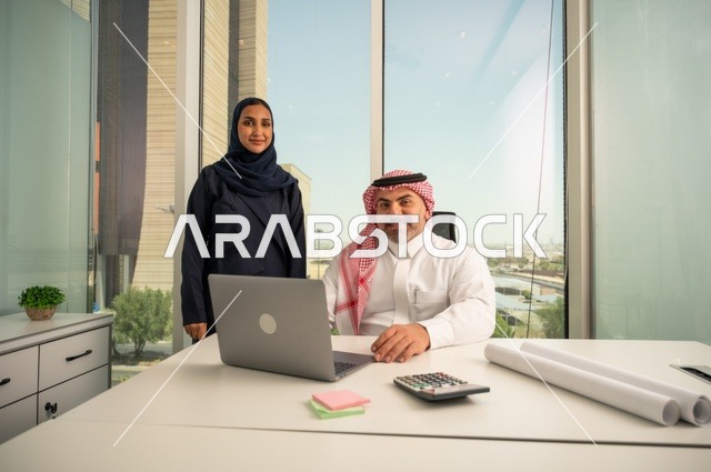 Looking at the camera with expressions of joy, using modern technology, an employee presenting projects to the manager for review via a laptop, studying and analyzing new projects, an Arab Gulf Saudi employee wearing traditional thobe and ghutrah sitting behind a desk inside the company headquarters, a Saudi office profession and job.