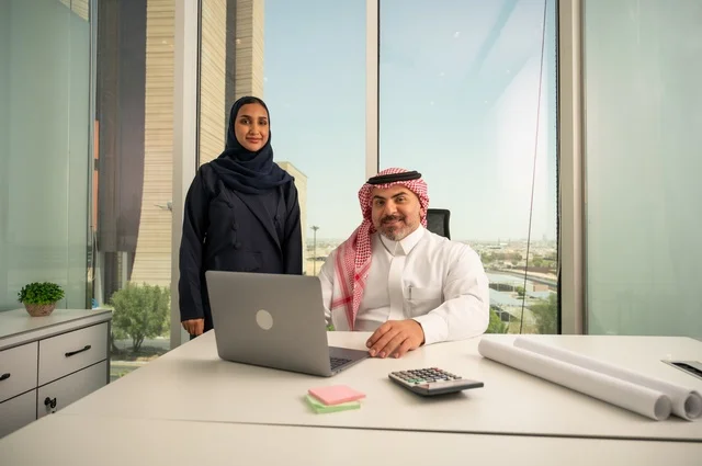 النظر إلى الكاميرا بتعابير السرور، استخدام جهاز تقني حديث، موظفة تقوم بتقديم المشاريع للمدير لتدقيقها عبر جهاز اللابتوب، دراسة وتحليل المشاريع الجديدة، موظف عربي خليجي سعودي يرتدي الثوب والشماغ التقليدي يجلس خلف المكتب داخل مقر الشركة، مهنة ووظيفة سعودية مكتبية