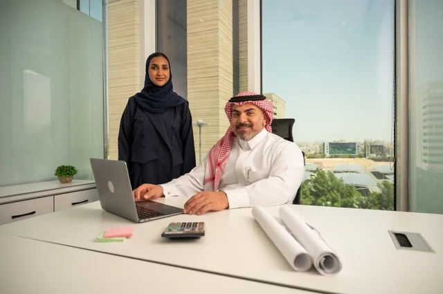 النظر إلى الكاميرا بتعابير السرور، استخدام جهاز تقني حديث، موظفة تقوم بتقديم المشاريع للمدير لتدقيقها عبر جهاز اللابتوب، دراسة وتحليل المشاريع الجديدة، موظف عربي خليجي سعودي يرتدي الثوب والشماغ التقليدي يجلس خلف المكتب داخل مقر الشركة، مهنة ووظيفة سعودية مكتبية