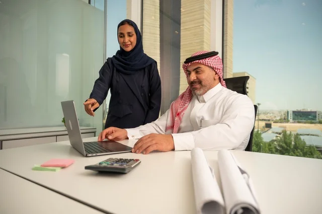 الوحدة والعمل الجماعي، استخدام جهاز تقني حديث، موظفة تقوم بتقديم المشاريع للمدير لتدقيقها عبر جهاز اللابتوب، تطوير ودراسة وتحليل المشاريع الجديدة، موظف عربي خليجي سعودي يرتدي الثوب والشماغ التقليدي يجلس خلف المكتب داخل مقر الشركة، مهنة ووظيفة سعودية مكتبية