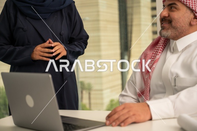 الوحدة والعمل الجماعي، استخدام جهاز تقني حديث، موظفة تقوم بتقديم المشاريع للمدير لتدقيقها عبر جهاز اللابتوب، تطوير ودراسة وتحليل المشاريع الجديدة، موظف عربي خليجي سعودي يرتدي الثوب والشماغ التقليدي يجلس خلف المكتب داخل مقر الشركة، مهنة ووظيفة سعودية مكتبية