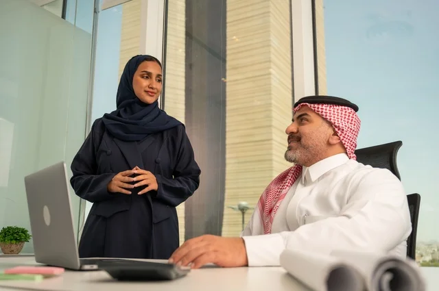 الوحدة والعمل الجماعي، استخدام جهاز تقني حديث، موظفة تقوم بتقديم المشاريع للمدير لتدقيقها عبر جهاز اللابتوب، تطوير ودراسة وتحليل المشاريع الجديدة، موظف عربي خليجي سعودي يرتدي الثوب والشماغ التقليدي يجلس خلف المكتب داخل مقر الشركة، مهنة ووظيفة سعودية مكتبية