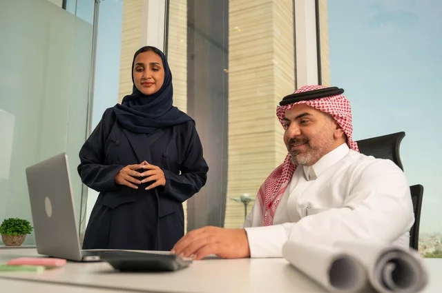 Unity and teamwork, using modern technical devices, an employee presenting projects to the manager for review via a laptop, developing, studying, and analyzing new projects, an Arab Gulf Saudi employee wearing traditional thobe and ghutrah sitting behind a desk inside the company headquarters, a Saudi office profession and job.