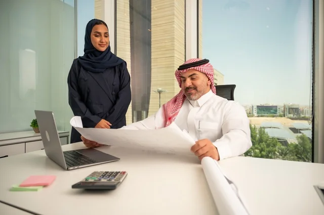Reviewing and reading plans, using modern technical devices, an employee presenting projects to the manager for review via a laptop, studying and analyzing new projects, an Arab Gulf Saudi employee wearing traditional thobe and ghutrah sitting behind a desk inside the company headquarters, a Saudi office profession and job.