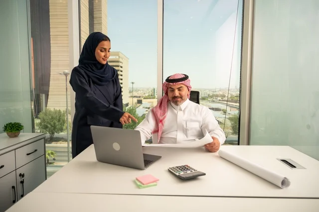 تدقيق وقراءة المخططات، استخدام جهاز تقني حديث، موظفة تقوم بتقديم المشاريع للمدير لتدقيقها عبر جهاز اللابتوب، دراسة وتحليل المشاريع الجديدة، موظف عربي خليجي سعودي يرتدي الثوب والشماغ التقليدي يجلس خلف المكتب داخل مقر الشركة، مهنة ووظيفة سعودية مكتبية