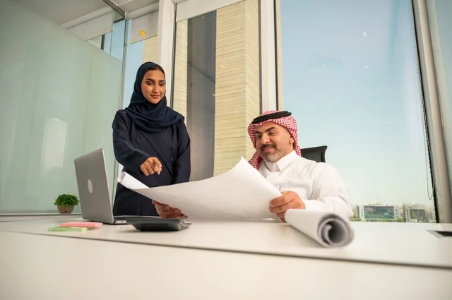 Reviewing and reading plans, using modern technical devices, an employee presenting projects to the manager for review via a laptop, studying and analyzing new projects, an Arab Gulf Saudi employee wearing traditional thobe and ghutrah sitting behind a desk inside the company headquarters, a Saudi office profession and job.