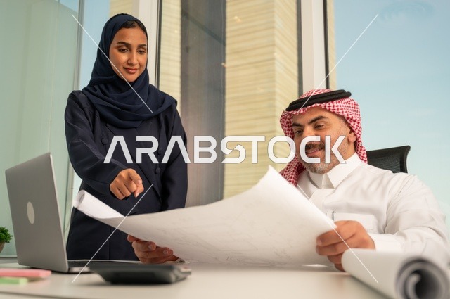 تدقيق وقراءة المخططات، استخدام جهاز تقني حديث، موظفة تقوم بتقديم المشاريع للمدير لتدقيقها عبر جهاز اللابتوب، دراسة وتحليل المشاريع الجديدة، موظف عربي خليجي سعودي يرتدي الثوب والشماغ التقليدي يجلس خلف المكتب داخل مقر الشركة، مهنة ووظيفة سعودية مكتبية