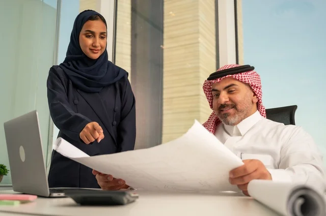 تدقيق وقراءة المخططات، استخدام جهاز تقني حديث، موظفة تقوم بتقديم المشاريع للمدير لتدقيقها عبر جهاز اللابتوب، دراسة وتحليل المشاريع الجديدة، موظف عربي خليجي سعودي يرتدي الثوب والشماغ التقليدي يجلس خلف المكتب داخل مقر الشركة، مهنة ووظيفة سعودية مكتبية