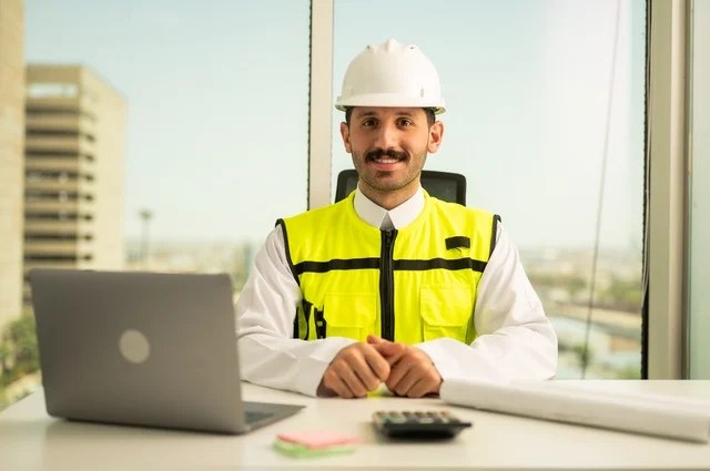 مهن ووظائف سعودية شبابية، أدوات هندسية لإكمال رسم مخطط البناء، دمج العمل بالتقنية والتكنولوجيا, رجل عربي خليجي سعودي مبتسم يرتدي الشماغ والثوب التقليدي يعمل على جهاز اللابتوب المحمول يجلس امام مكتب عليه خوذة الحماية والمخططات، تدقيق المشاريع ومراجعتها