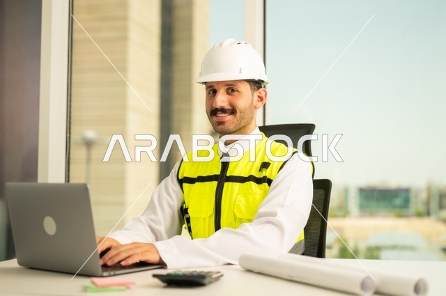 Project auditing and review, engineering tools for completing building plan drawings, integrating work with technology and tech, an Arab Gulf Saudi man wearing a ghutrah and traditional thobe working on a laptop sitting in front of a desk with a safety helmet and plans.