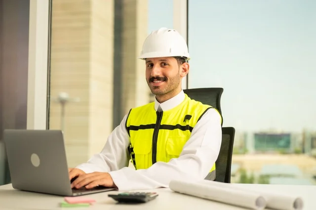 Project auditing and review, engineering tools for completing building plan drawings, integrating work with technology and tech, an Arab Gulf Saudi man wearing a ghutrah and traditional thobe working on a laptop sitting in front of a desk with a safety helmet and plans.