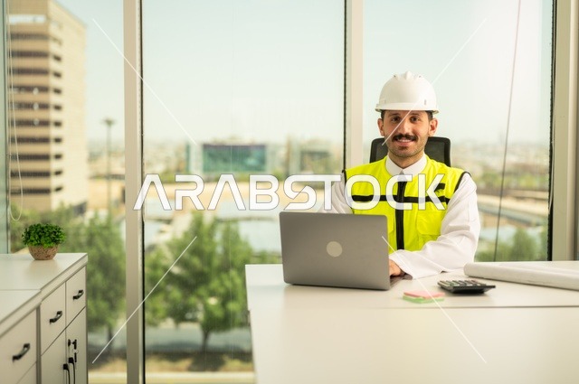 Project auditing and review, engineering tools for completing building plan drawings, integrating work with technology and tech, an Arab Gulf Saudi man wearing a ghutrah and traditional thobe working on a laptop sitting in front of a desk with a safety helmet and plans.