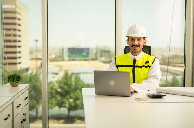 Project auditing and review, engineering tools for completing building plan drawings, integrating work with technology and tech, an Arab Gulf Saudi man wearing a ghutrah and traditional thobe working on a laptop sitting in front of a desk with a safety helmet and plans.