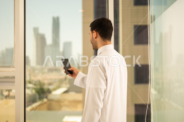 Using a modern and advanced technical device, a close-up image from the back of a Saudi Arabian Gulf man wearing traditional thobe working on a mobile phone, progress and architectural development in the Gulf countries, a background of towers and skyscrapers in the city of Riyadh