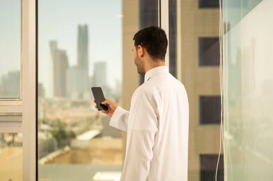 Using a modern and advanced technical device, a close-up image from the back of a Saudi Arabian Gulf man wearing traditional thobe working on a mobile phone, progress and architectural development in the Gulf countries, a background of towers and skyscrapers in the city of Riyadh