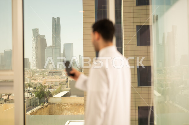 استخدام جهاز تقني حديث ومتطور،  صورة مقربة من الخلف لرجل عربي خليجي سعودي يرتدي الثوب التقليدي يعمل على الهاتف المحمول، التقدم والتطور المعماري في دول الخليج، خلفية الأبراج وناطحات السحاب في مدينة الرياض